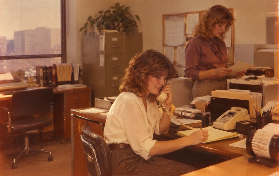 Two women in an office with desks, papers, and a rotary phone. One woman is seated, talking on the phone and writing, while the other stands reading a document. Sunlight comes through a window.