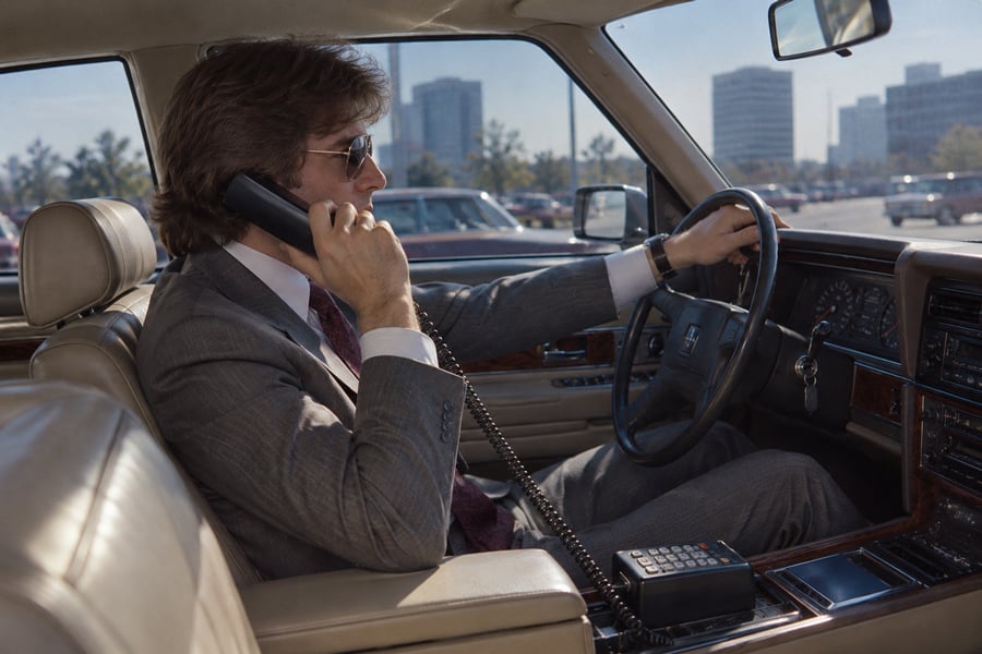 A man in a gray suit drives a car while talking on a large car phone, with city buildings visible through the window on a sunny day.