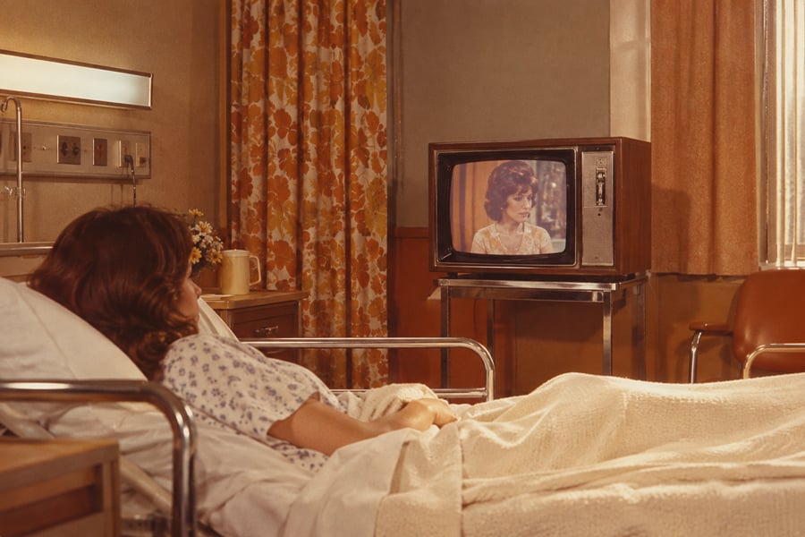 A woman in a hospital bed watches a television showing a woman speaking. The hospital room has floral curtains, medical equipment, and warm lighting.