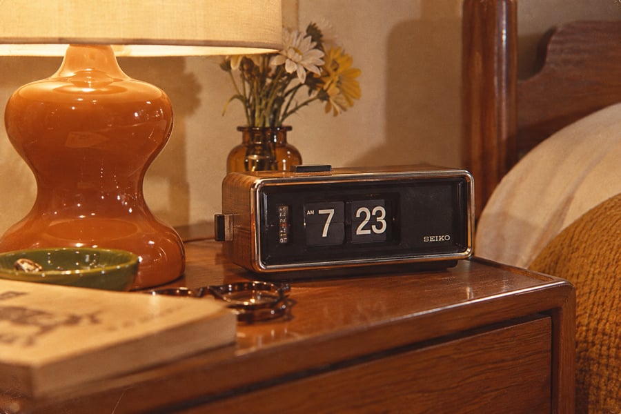 A vintage flip clock reading 7:23 sits on a wooden nightstand beside a lamp, small vase with flowers, green dish, eyeglasses, and a book. A mustard colored blanket is visible on the bed next to it.
