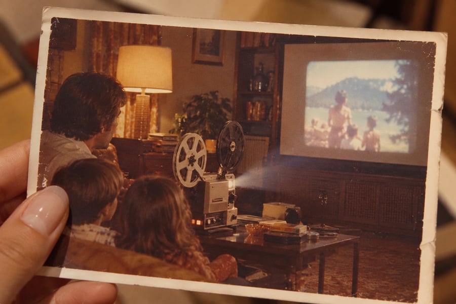 A hand holds an old photo showing a family watching a home movie projected onto a screen in a cozy living room, with a vintage film projector on the table.