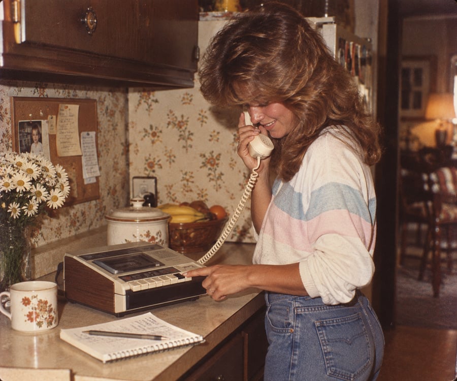 A woman with long hair stands in a kitchen, smiling while using a corded landline phone. She wears a striped shirt and jeans, with a notepad, pen, mug, and flowers on the counter beside her.