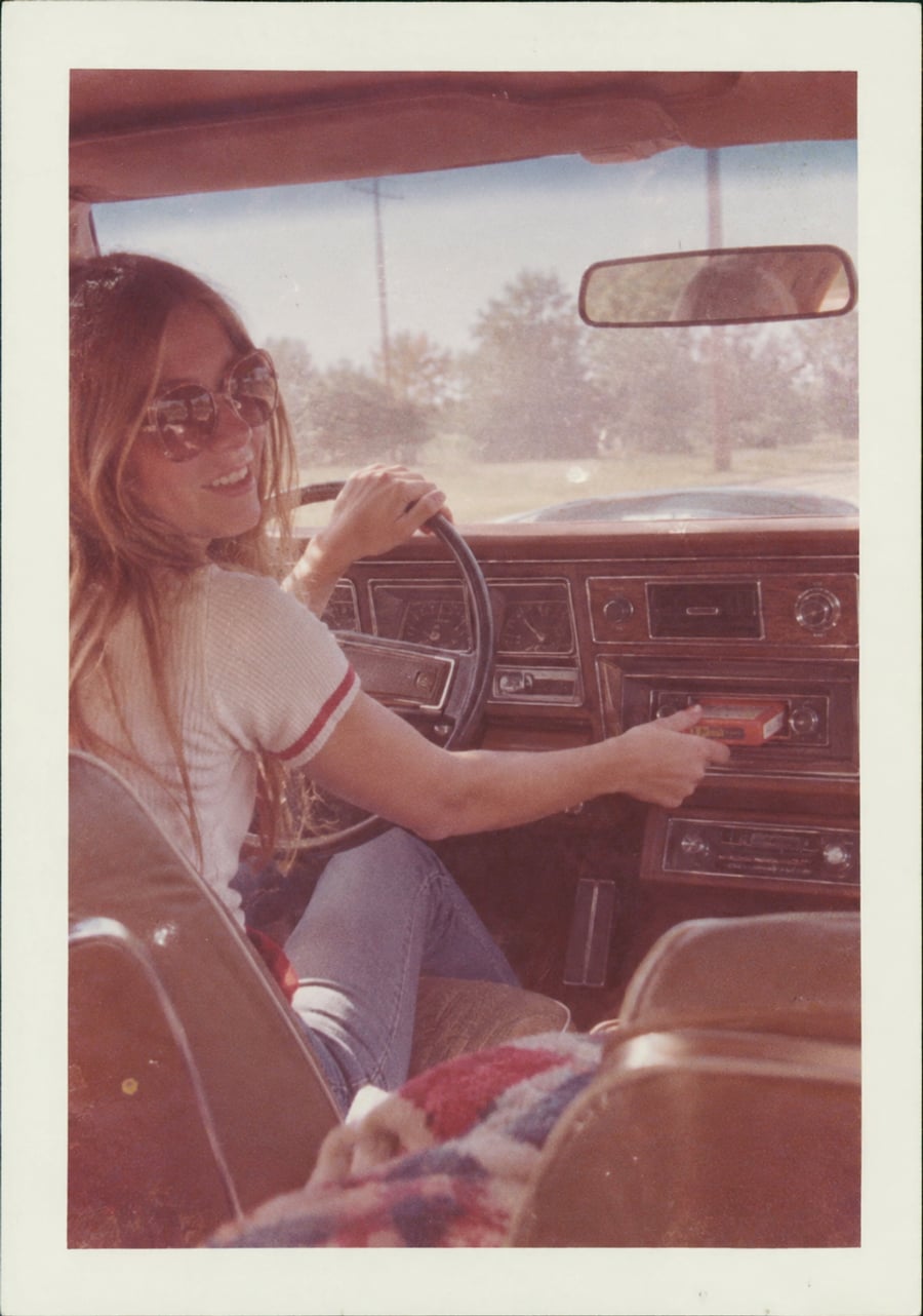 A woman with long hair and sunglasses smiles while sitting in the driver’s seat of a vintage car, holding a cassette tape near the dashboard. Sunlight streams in from the window, and trees are visible outside.