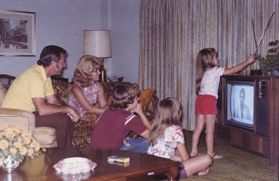 A family sits in a living room watching TV; a girl stands adjusting the antenna while the others sit on chairs and the floor, watching her.