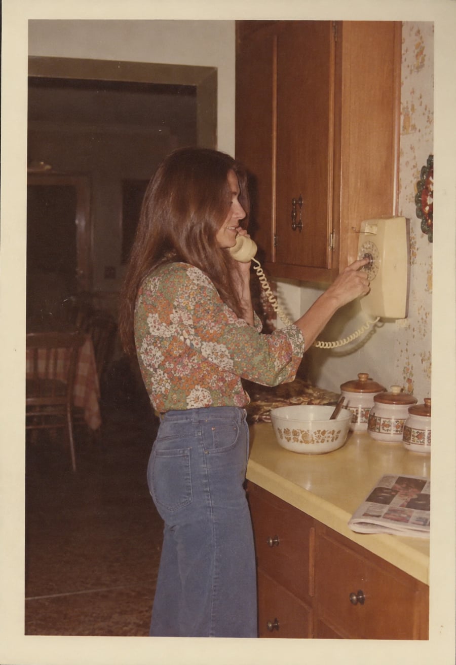 A woman with long hair and a floral shirt uses a yellow rotary phone in a vintage kitchen, standing by a yellow counter with mixing bowls, canisters, and a newspaper.