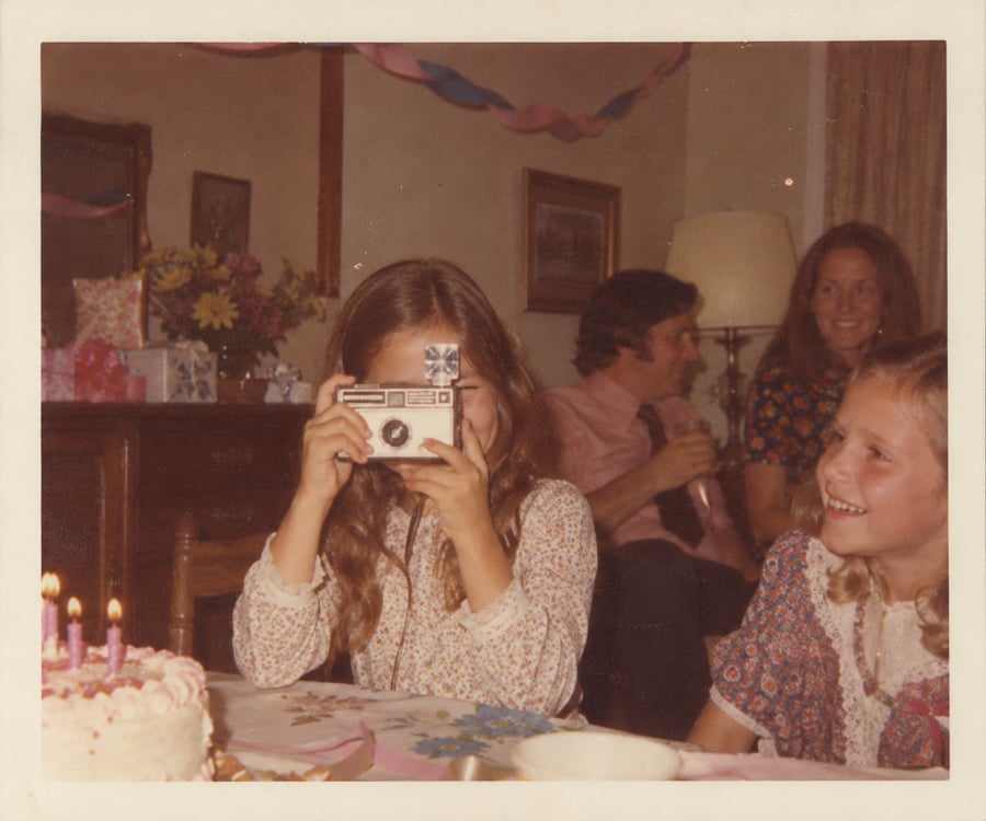A girl takes a photo with a vintage camera at a birthday party. Another girl smiles beside her. Adults and party decorations, including a cake and streamers, are visible in the background.