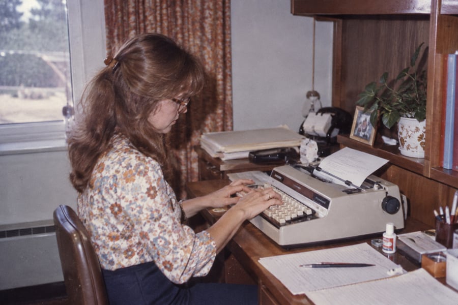 A woman with long hair and glasses types on a typewriter at a desk with papers, a plant, books, and a phone. A window with curtains is in the background.