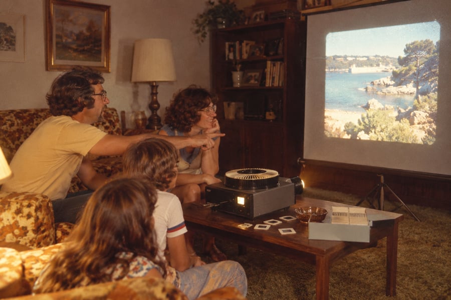 Four people sit on a couch in a living room, watching vacation slides projected onto a screen. A slide projector and photo slides are on the coffee table in front of them.