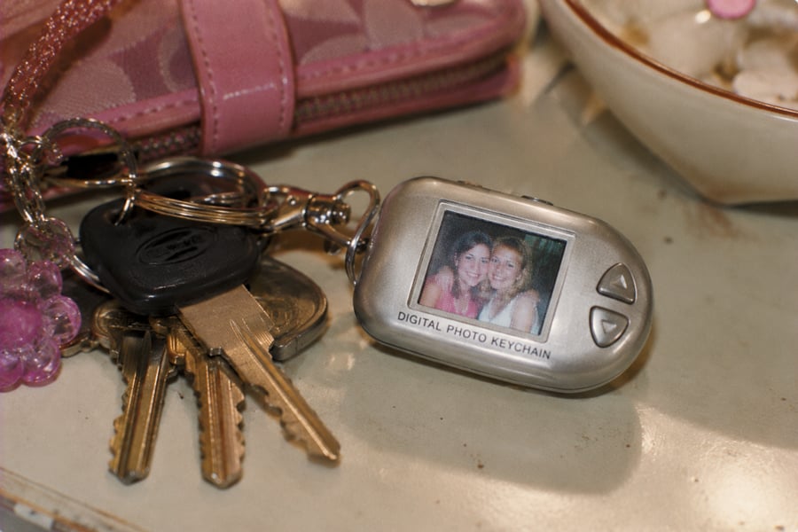 A set of keys with a digital photo keychain displaying a picture of two smiling people, next to a pink wallet and a cream-colored dish.