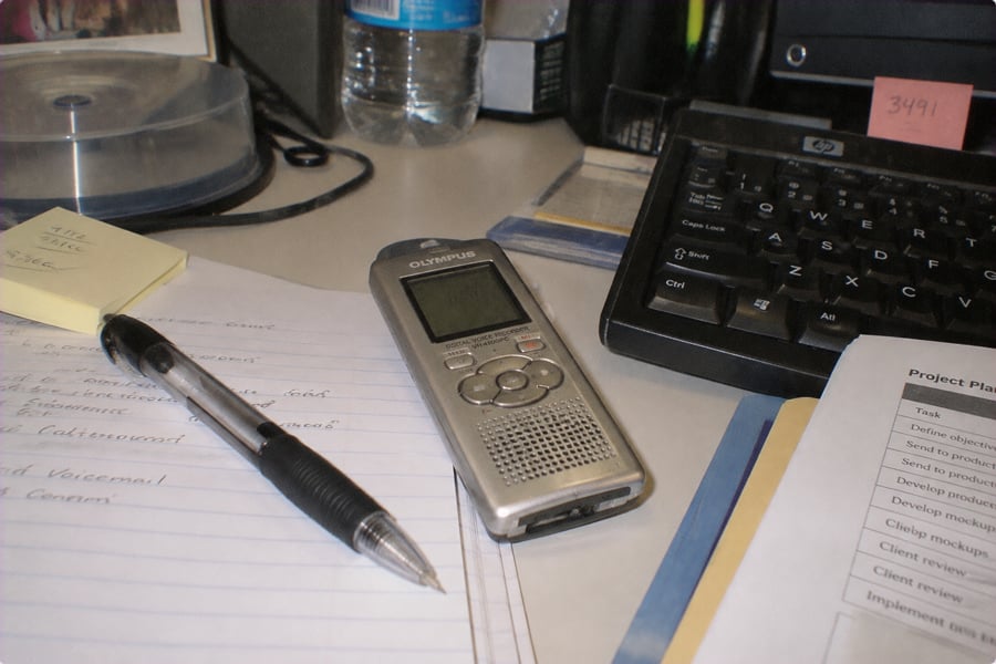 A digital voice recorder, pen, and papers are on a cluttered office desk with a keyboard, water bottle, sticky notes, and project plan visible.