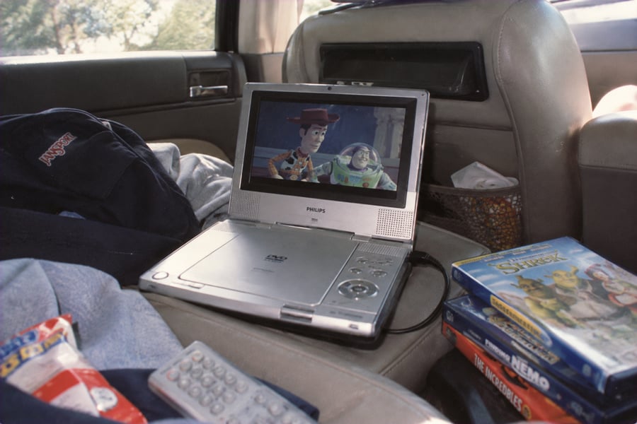 A portable DVD player shows a scene from Toy Story inside a car. Nearby are DVDs for Shrek and Finding Nemo, snacks, a remote, and a jacket on the seat.
