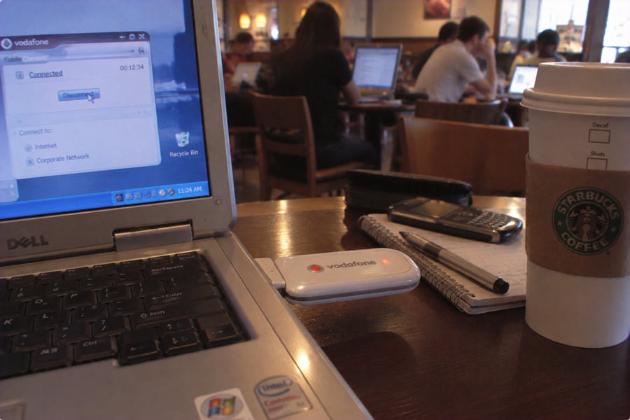 A Dell laptop, Vodafone USB modem, phone, and a Starbucks coffee cup sit on a table in a busy cafe with several people working on laptops in the background.