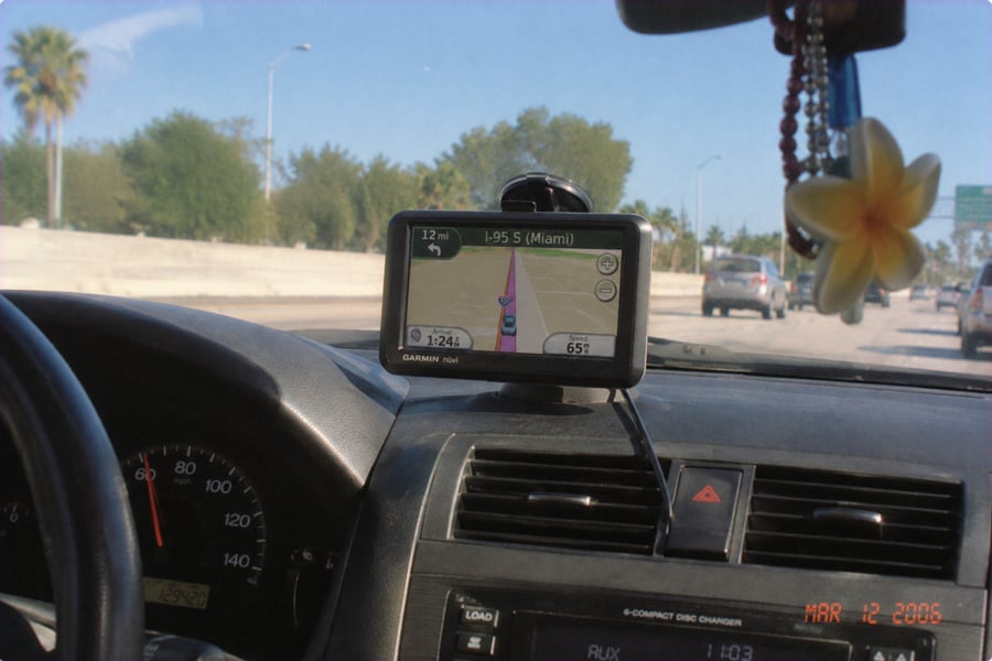 A GPS device shows directions to Miami on a car dashboard. The highway is visible through the windshield, and cars are ahead. A flower charm hangs from the rearview mirror. The date on the photo is March 12, 2006.
