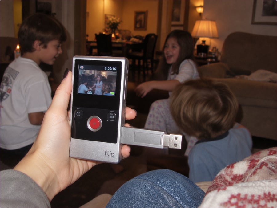 A hand holds a Flip video camera showing a recording of three children laughing and playing in a cozy living room.