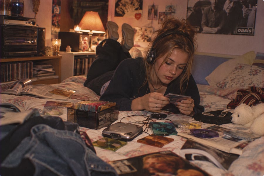 A teenage girl with headphones lies on her bed, looking at a CD. Her room is cluttered with CDs, magazines, a Discman, and posters. Warm, dim lighting creates a cozy, nostalgic atmosphere.