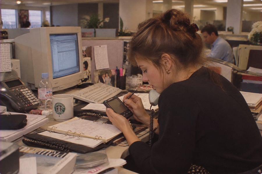 A woman sits at a cluttered office desk, talking on a corded phone while using a PDA. Papers, a computer, calendar, phone, and coffee mug are on the desk. Other people work in the background.