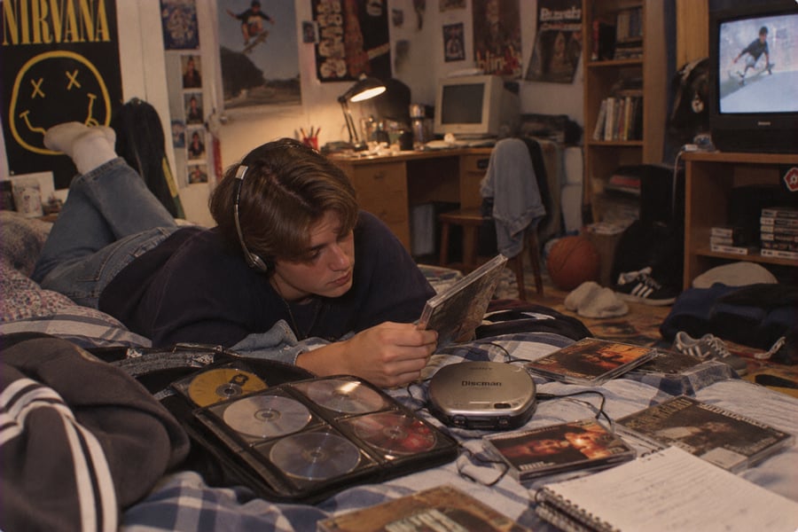 Teen wearing headphones lies on a bed, looking at a CD booklet. A portable CD player, CDs, and a notebook are nearby. The room has posters, a TV, and cluttered shelves, creating a cozy, retro atmosphere.