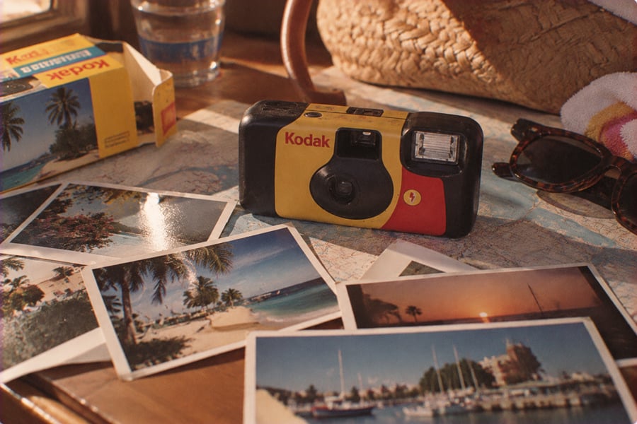 A Kodak disposable camera sits on a table with several travel photos, a map, sunglasses, a towel, and a straw bag nearby, suggesting a vacation scene.