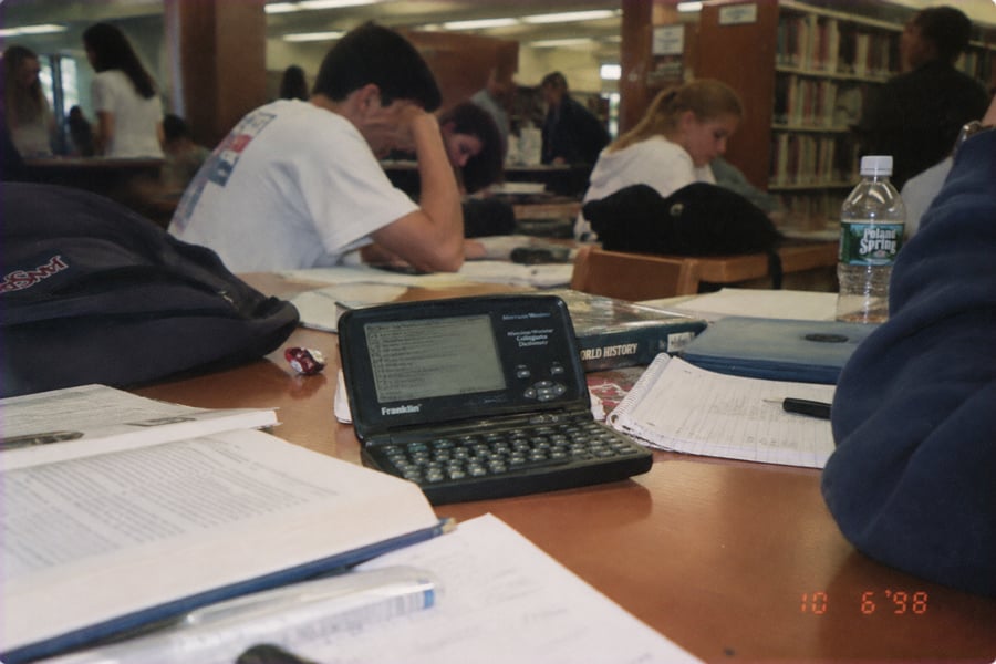 Students study at a library table with open books, papers, a Franklin electronic dictionary, water bottle, and backpacks visible. The scene appears to be from the 1990s based on the technology and date stamp.