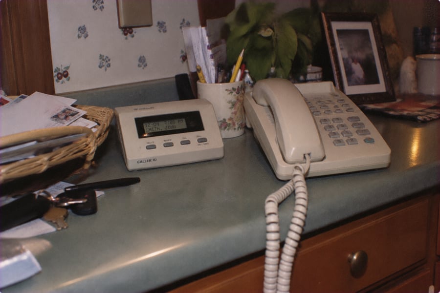 A beige corded landline phone and caller ID device sit on a green countertop surrounded by papers, keys, a mug with pens, and a framed photo in a home setting.
