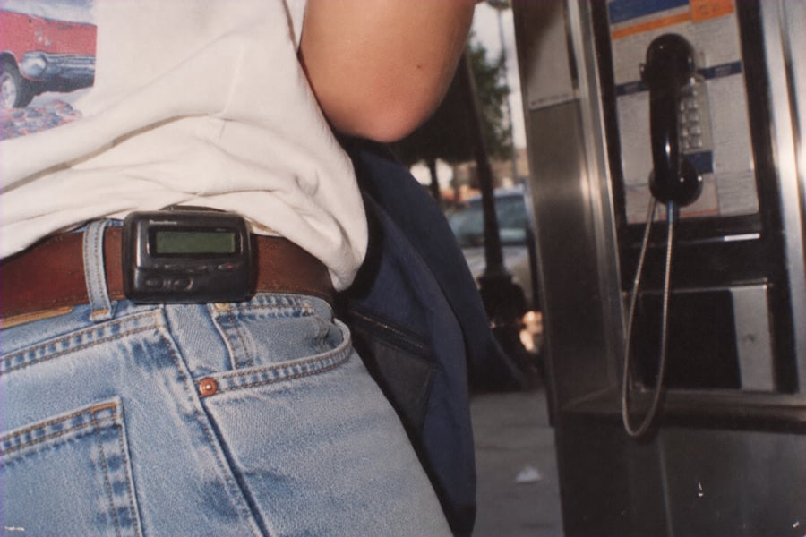 A person wearing jeans and a white shirt with a pager clipped to their belt stands near a public payphone. The scene appears to be outdoors.