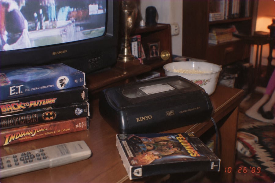 A stack of VHS tapes, a VCR, a bowl of popcorn, and a TV playing a movie sit on a wooden table. Visible tapes include E.T., Back to the Future, Batman, Indiana Jones, and The Goonies.