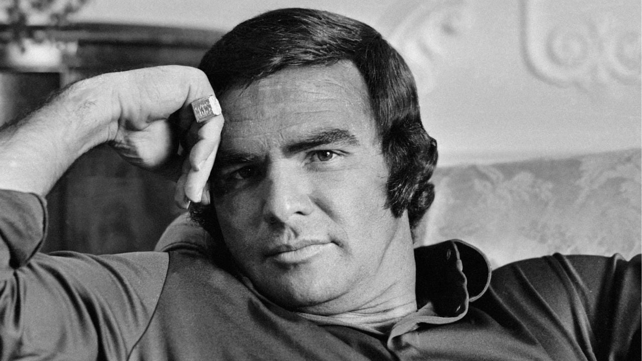 A man with short dark hair and sideburns rests his head on his hand, wearing a ring and a collared shirt, looking directly at the camera with a neutral expression. The image is in black and white.