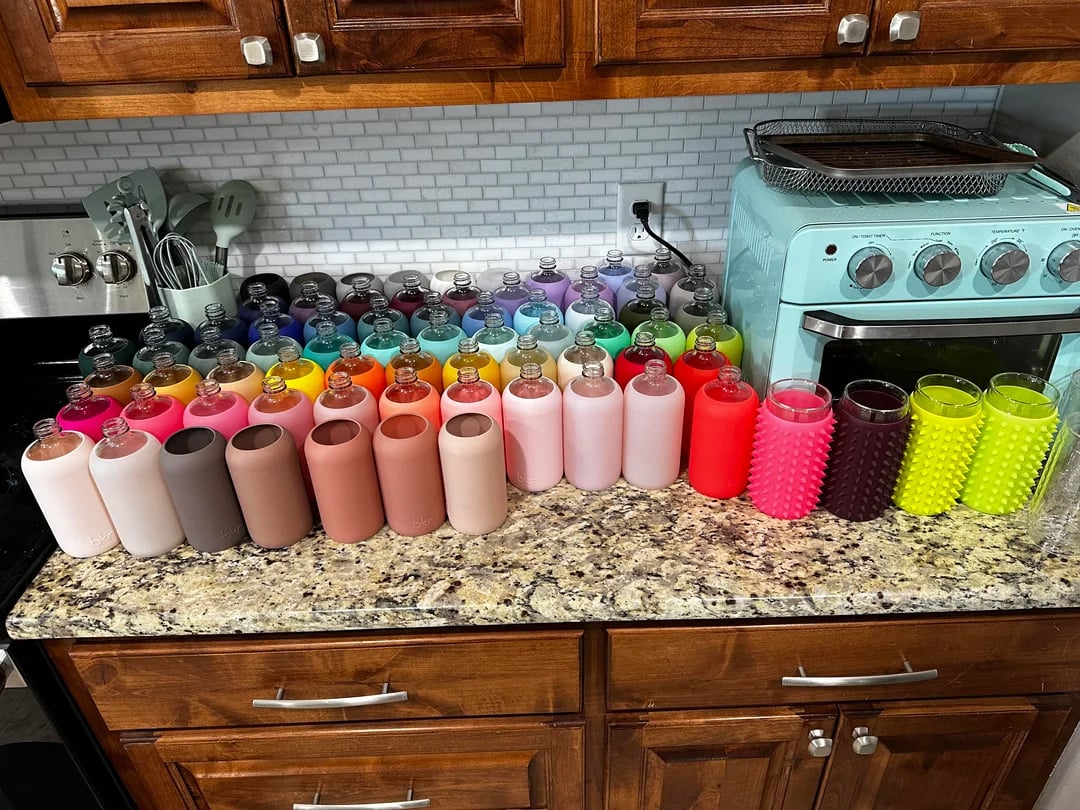 Dozens of colorful glass water bottles with silicone sleeves are arranged on a kitchen countertop in neat rows beneath wooden cabinets. A mint green oven and kitchen tools are visible in the background.