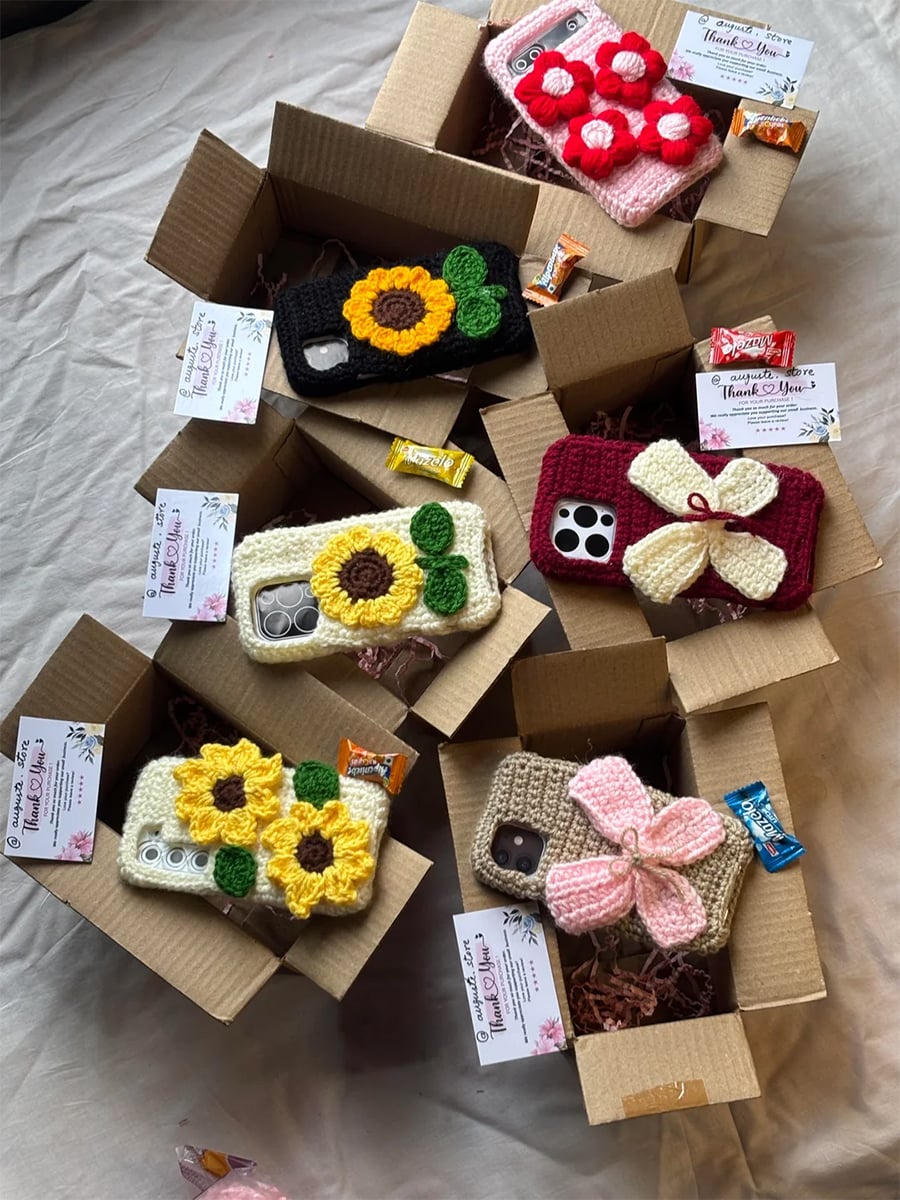 Six cardboard boxes contain crocheted phone cases with flower designs, business cards, and candies, all arranged on a beige fabric background. Cases are various colors including yellow, pink, red, green, and brown.