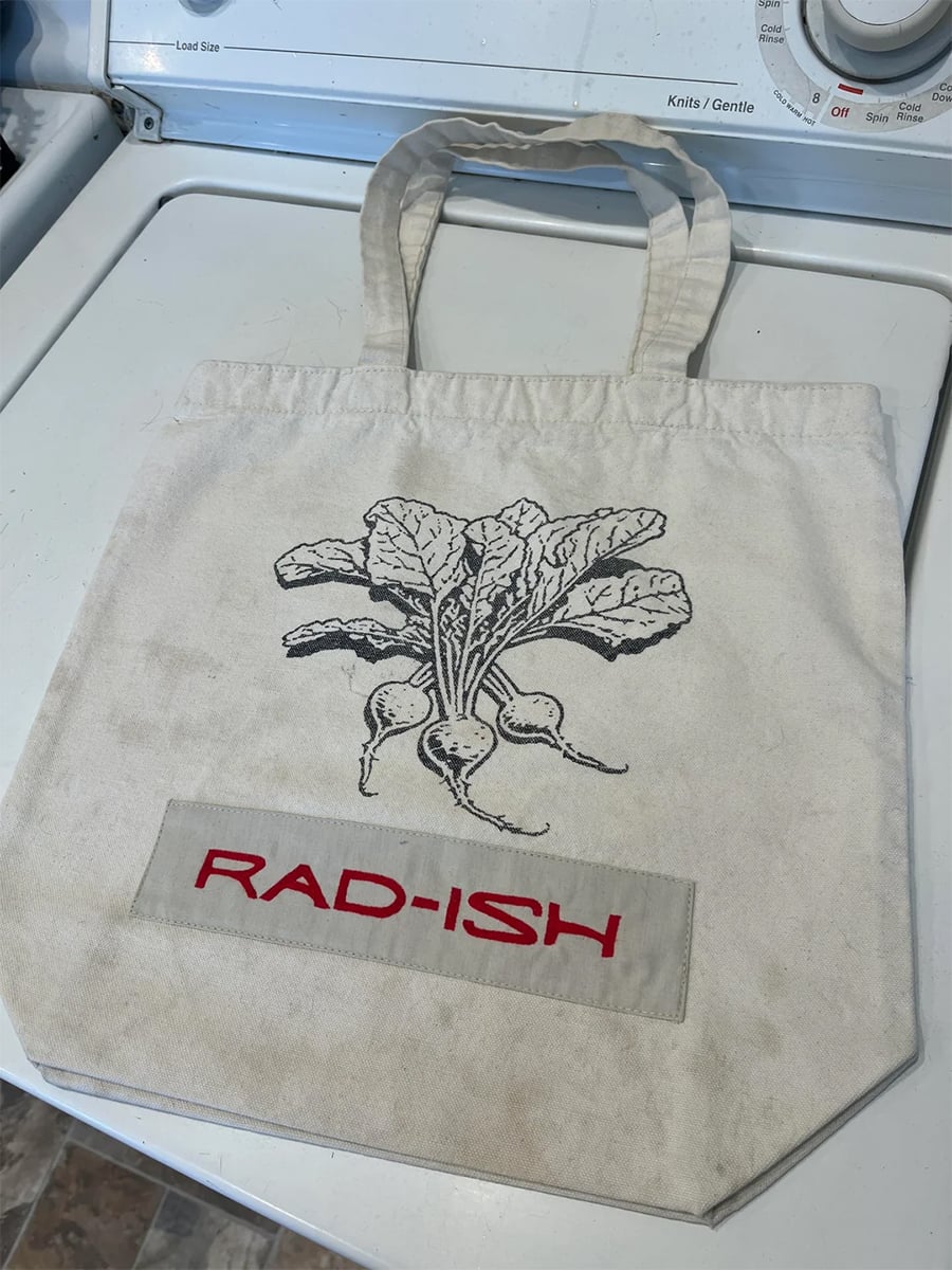 A canvas tote bag with a black radish illustration and the word “RAD-ISH” in bold red letters, sitting on a white washing machine.