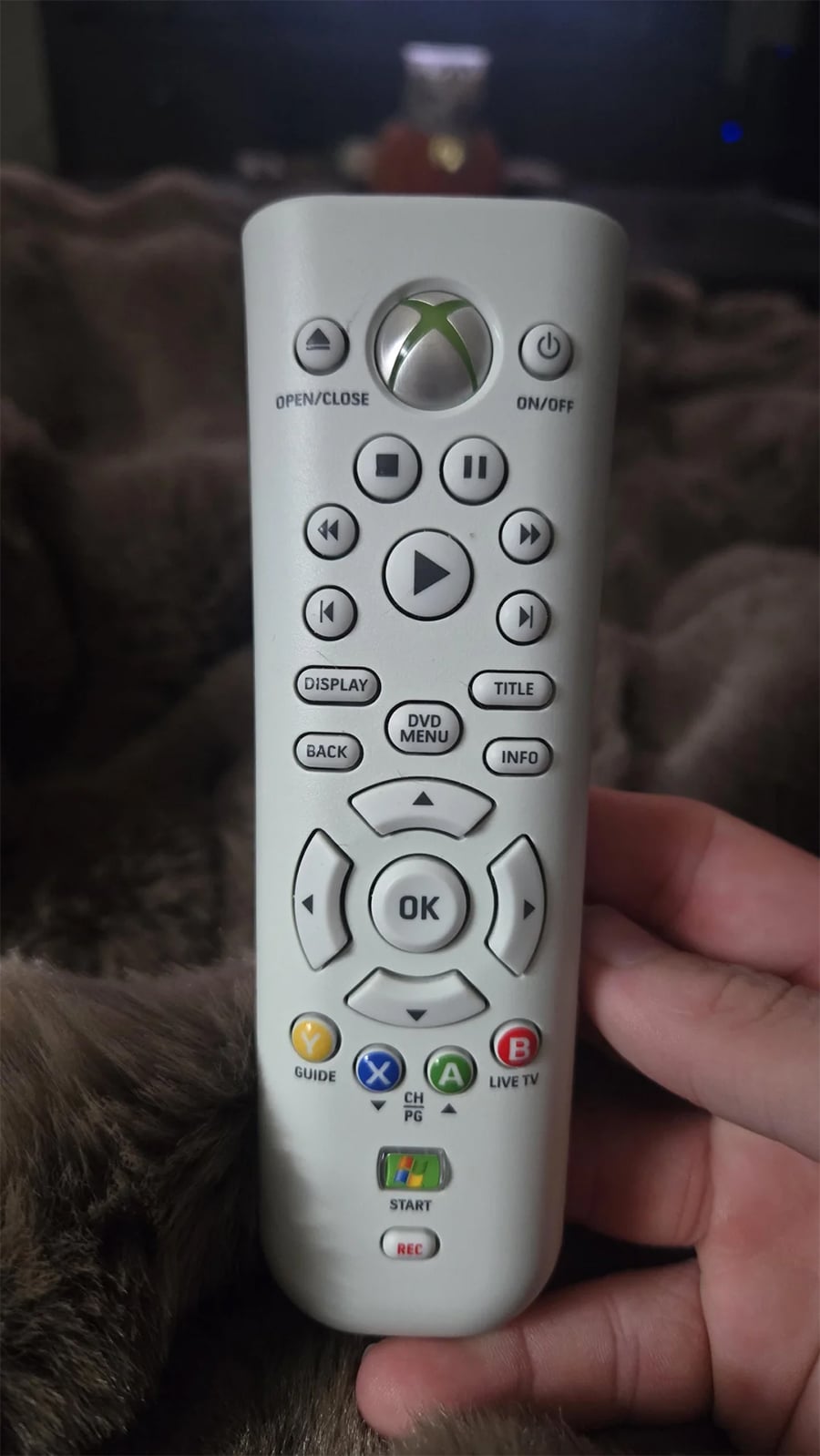 A hand holding a white Xbox remote control with various buttons, including play, pause, directional arrows, and colored A, B, X, Y buttons. A TV and blanket are visible in the background.