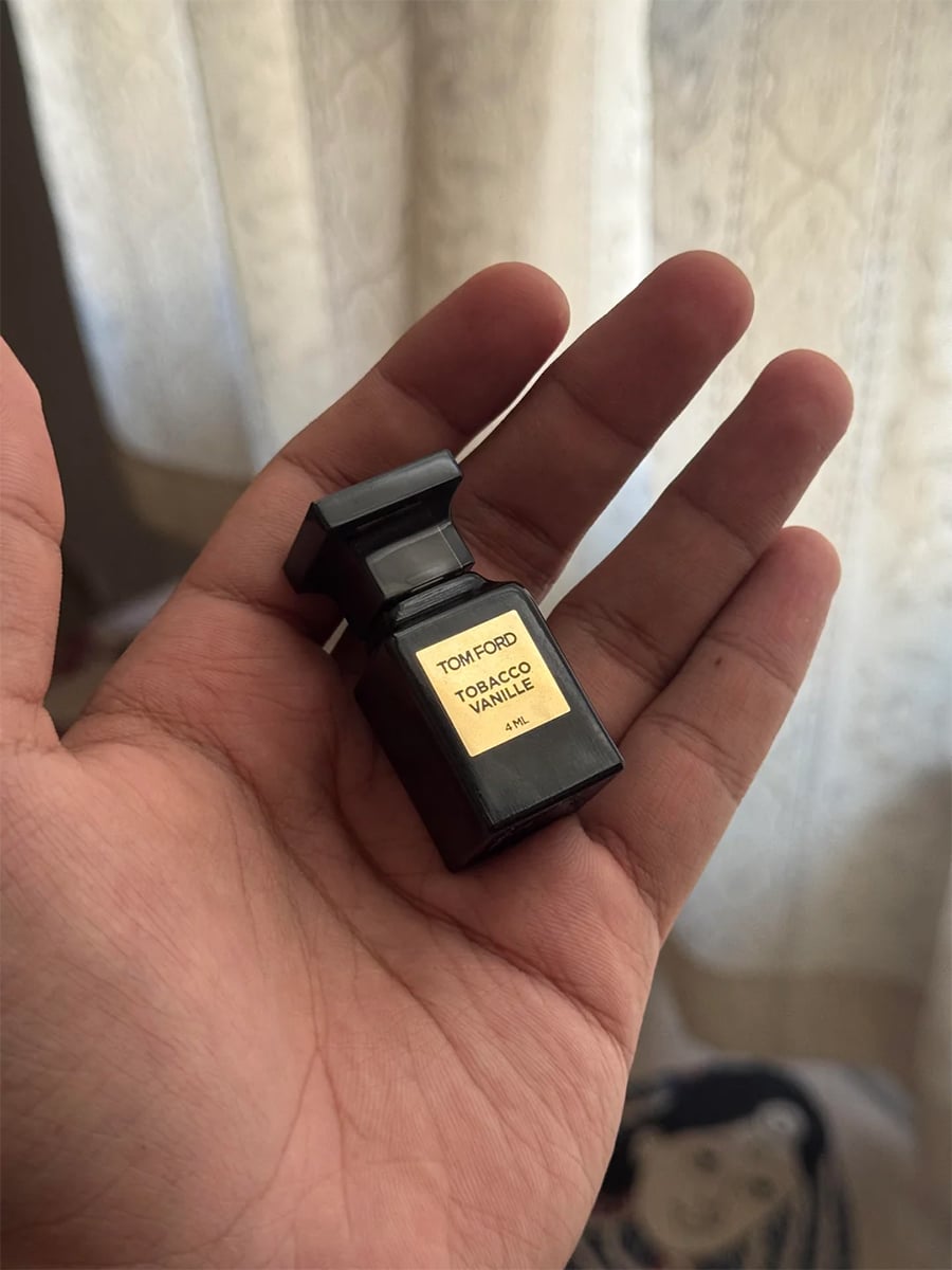 A hand holds a small black bottle of Tom Ford Tobacco Vanille perfume with a gold label, against a background of sheer curtains.