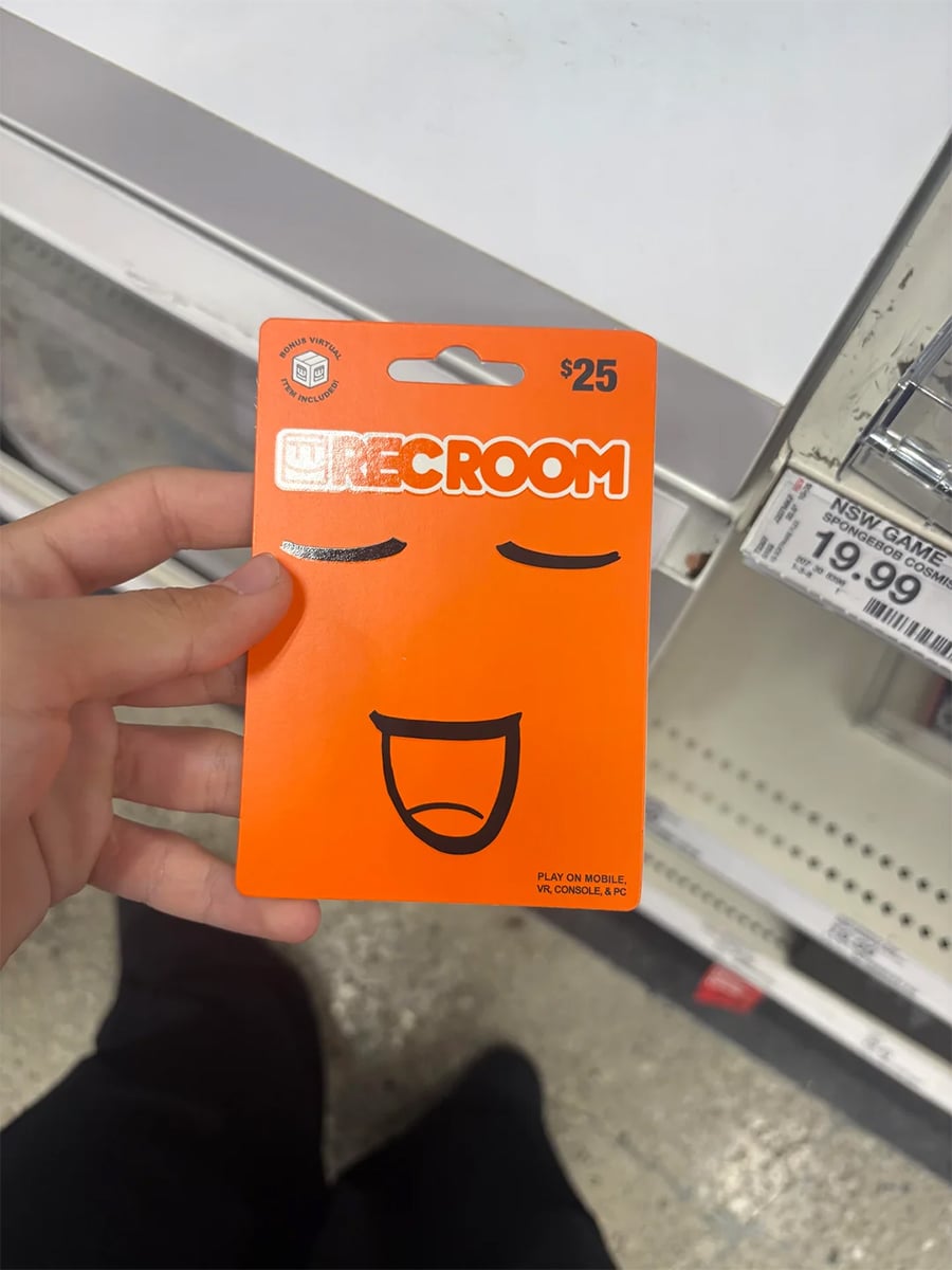 A hand holds a $25 Rec Room gift card with a smiling face design in a store aisle. A price tag nearby reads $19.99. The card is orange with closed eyes and an open-mouthed smile.