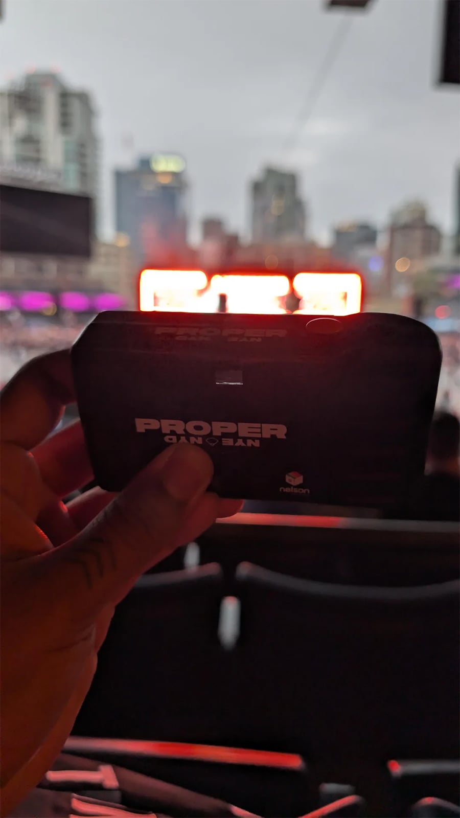 A hand holds a small black object labeled “PROPER CAN U SAN” with a blurred stadium and bright scoreboard in the background.