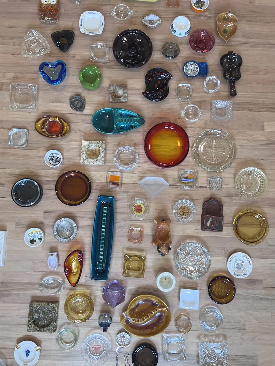 Assorted vintage ashtrays of various shapes, sizes, and colors are neatly arranged on a light wood floor, viewed from above.