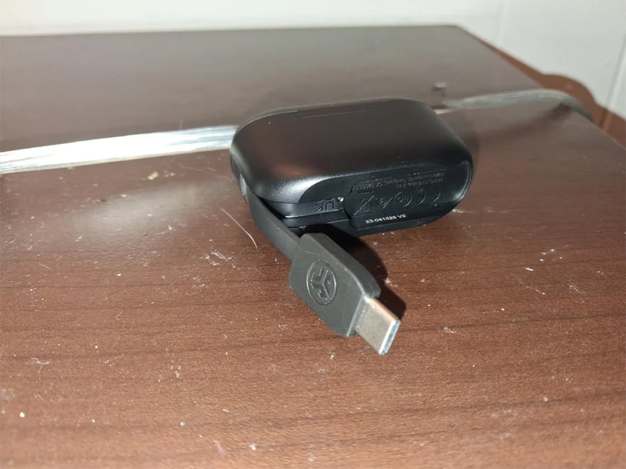 A black wireless earbud case with a USB-C charging cable attached rests on a brown wooden surface under bright light.