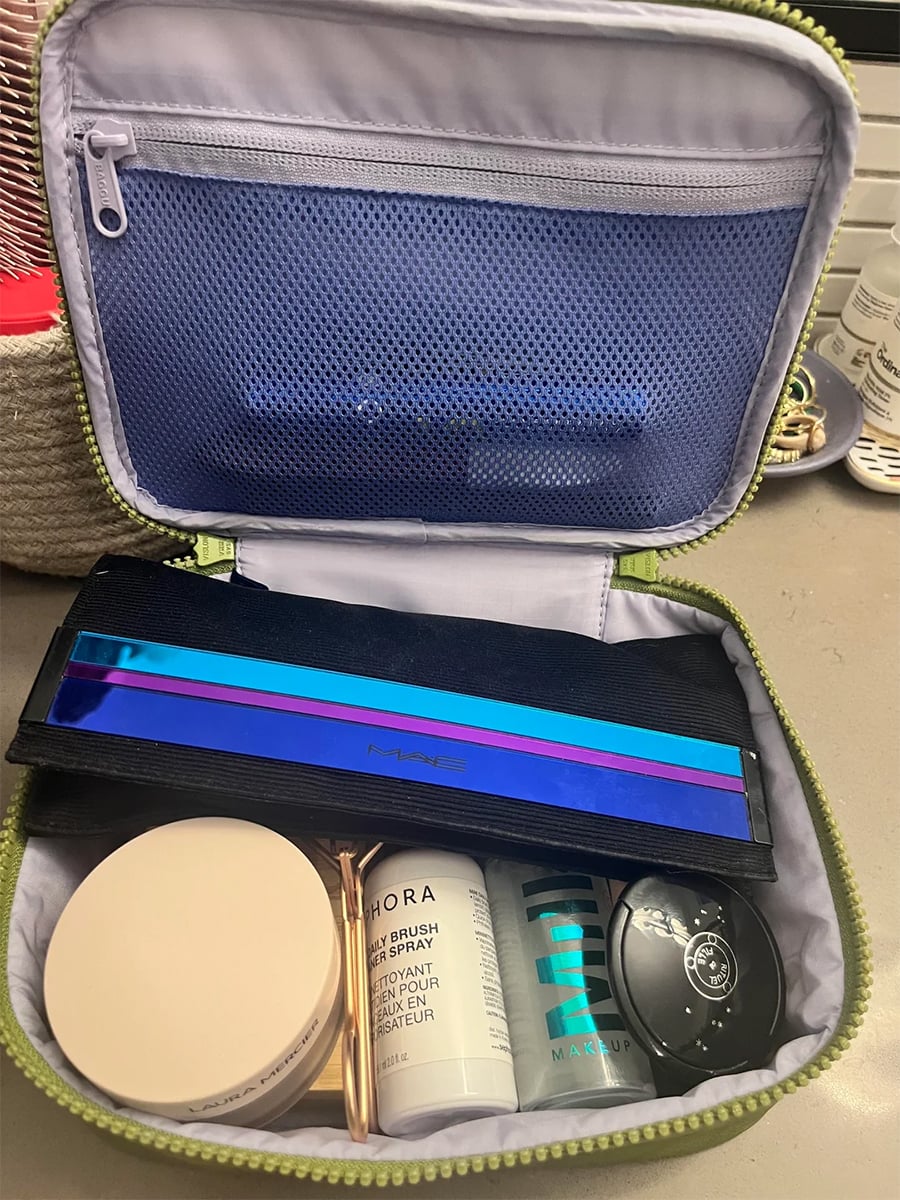 Open makeup bag showing a palette, round compact, brushes, and bottles of setting spray and makeup, all organized neatly inside a green zippered case on a countertop.