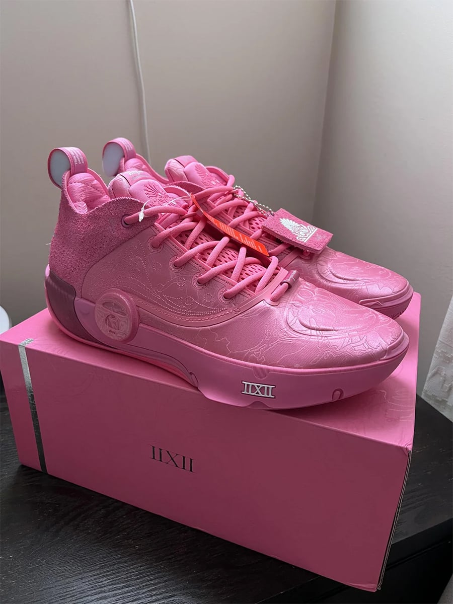 Pair of bright pink athletic shoes with textured patterns, sitting on a matching pink shoe box with the Roman numerals “IIZII” printed on the front, positioned on a dark wood surface near a white wall.
