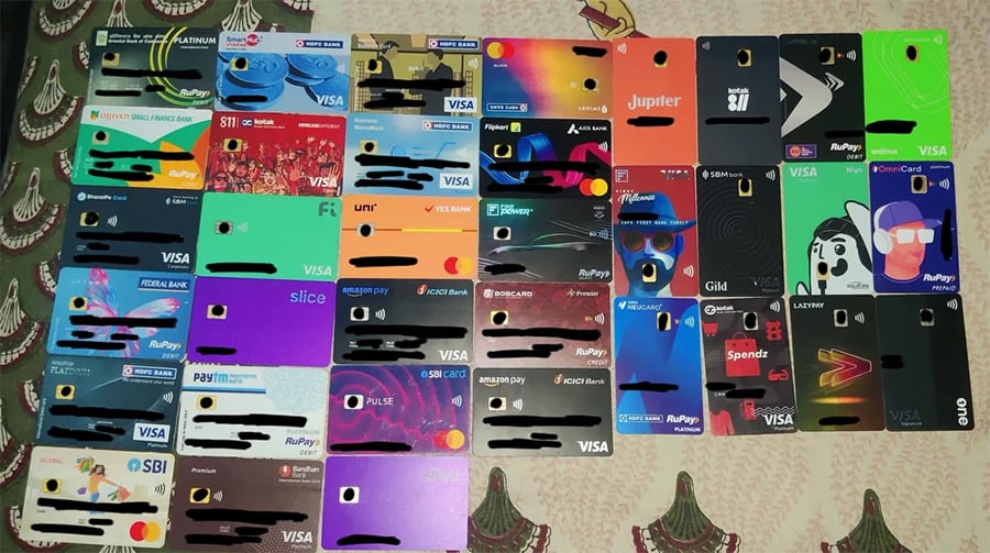 A collection of 35 colorful credit and debit cards from various banks and financial institutions is arranged in a grid on a patterned surface. Card numbers and names are blacked out for privacy.