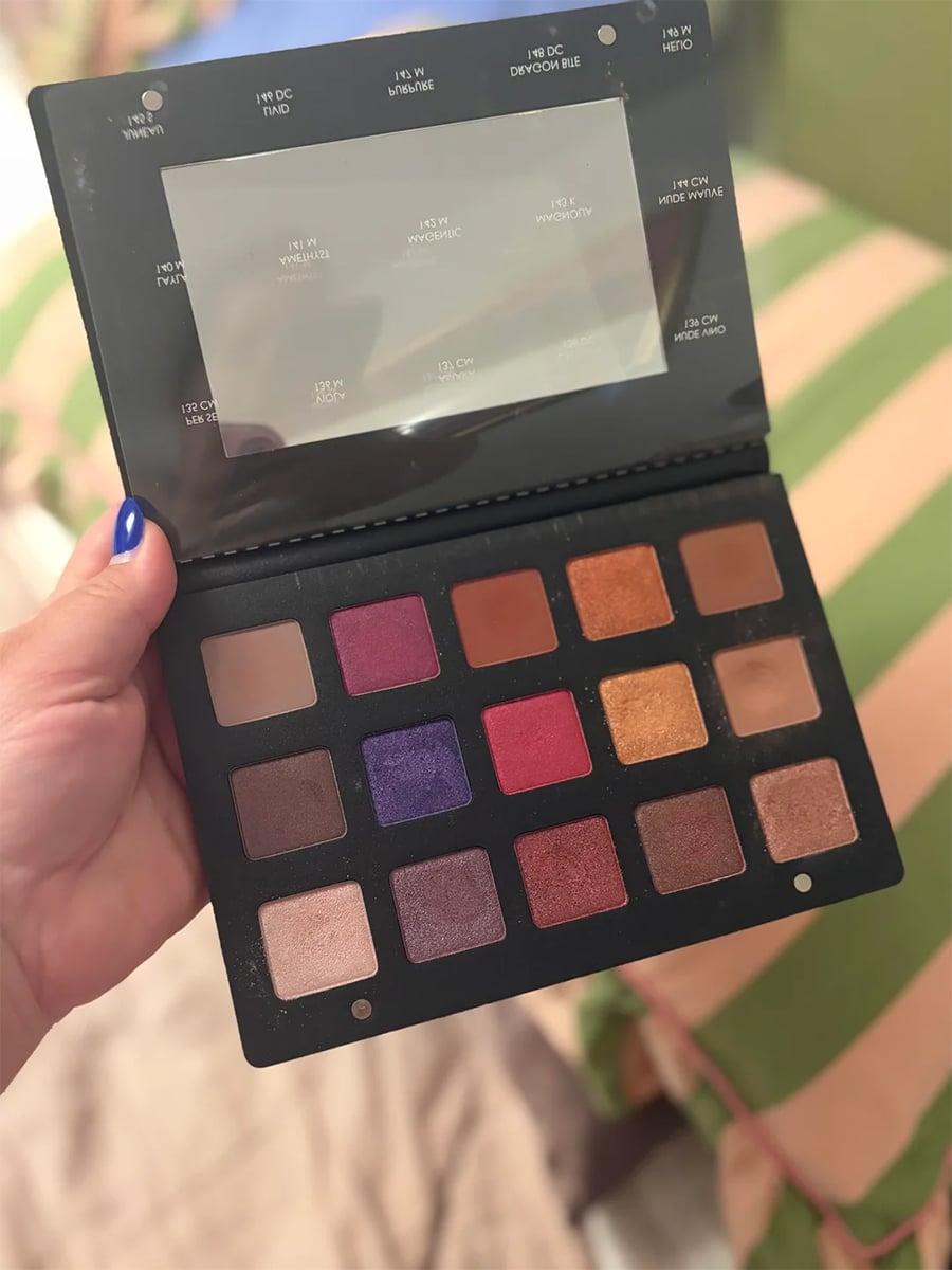 A hand holding an open eyeshadow palette with 15 colorful shades, including gold, red, purple, and brown, in front of a striped green and pink background.