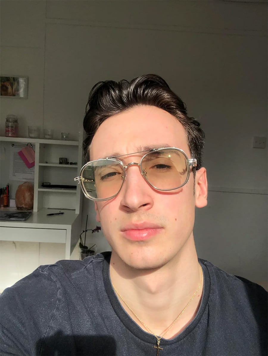 A person with short dark hair and large clear glasses takes a selfie in bright sunlight, wearing a dark shirt and a gold necklace. A white desk and shelves with various items are visible in the background.