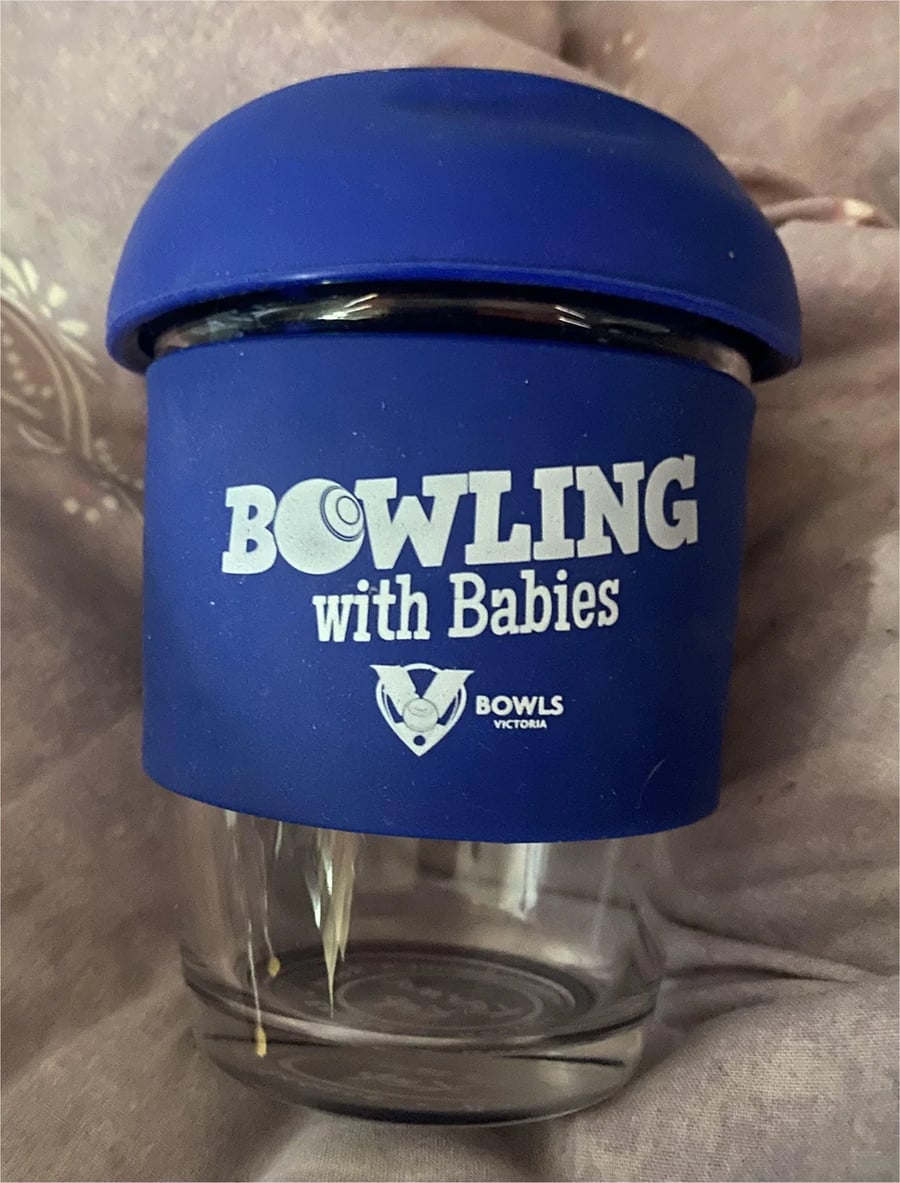 A glass cup with a blue silicone lid and sleeve that reads “Bowling with Babies” above the Bowls Victoria logo, photographed against a soft, pinkish background.