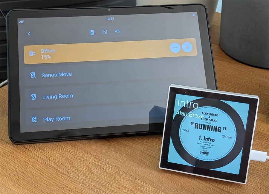 A tablet displays smart home controls, and a square digital device shows the song “Running” by Alan Braxe & Fred Falke. Both sit on a wooden surface next to a gray pot.