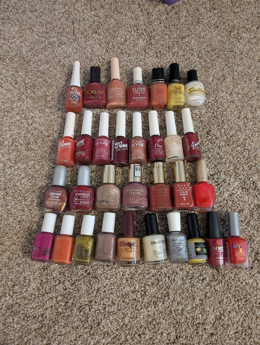 Thirty bottles of nail polish in various colors and brands are neatly arranged in rows on a light brown carpeted floor. The colors range from reds and pinks to gold, silver, orange, and clear.