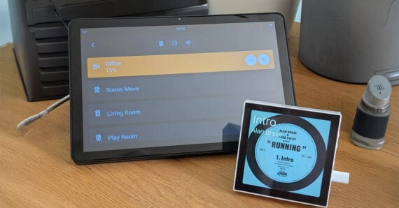 A tablet shows a home audio control screen on a desk next to a small smart display displaying music info, a pen, and a gray canister.