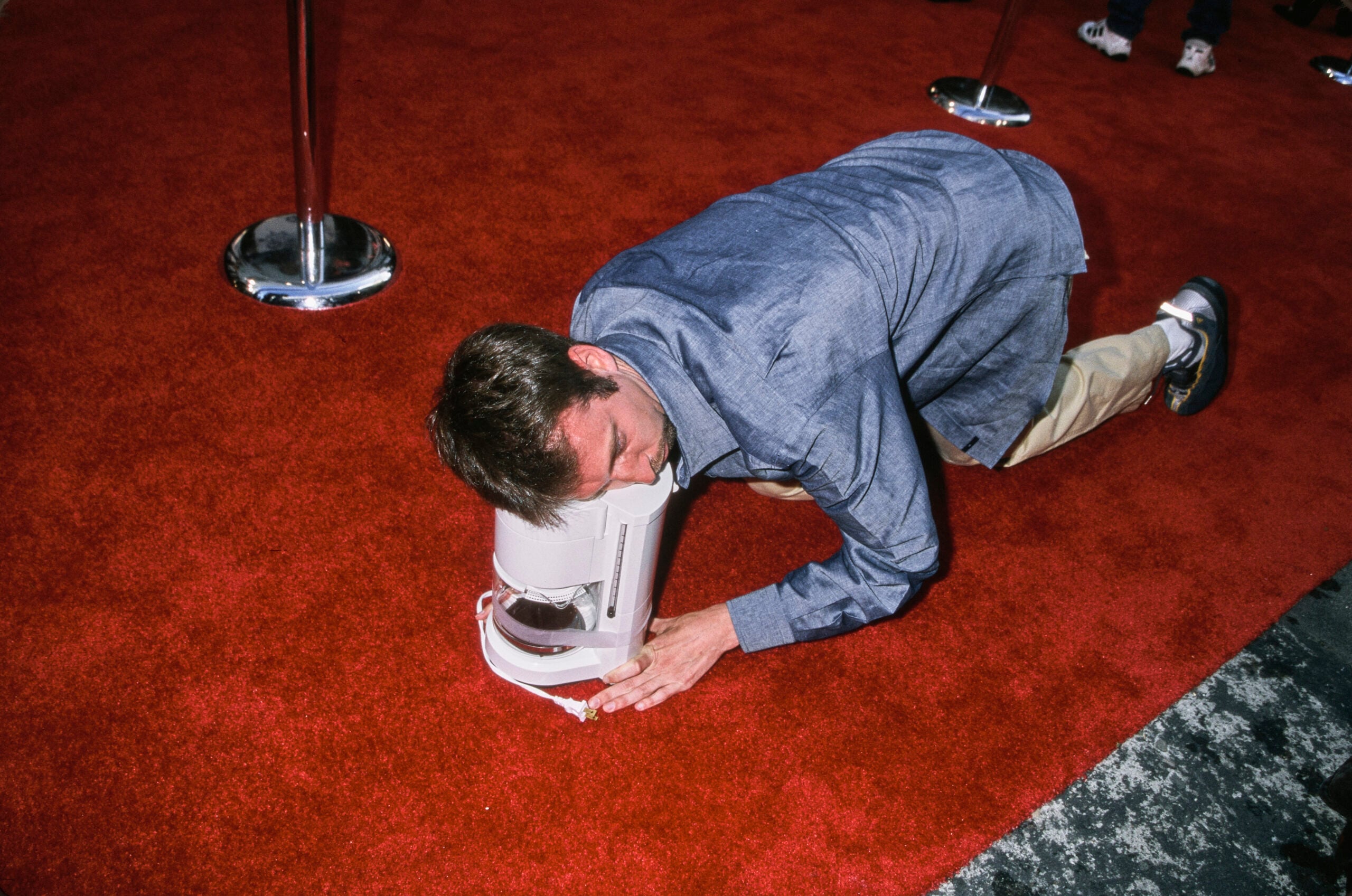 A man in a blue shirt kneels on a red carpet with his head inside a white machine, appearing to interact with or examine it closely.