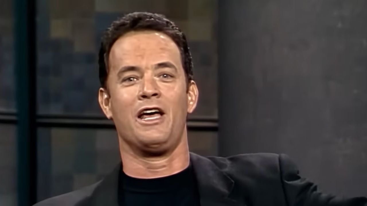 A man with short dark hair wearing a black shirt and jacket is speaking animatedly, with his mouth open and arm raised, against a blurred indoor background.