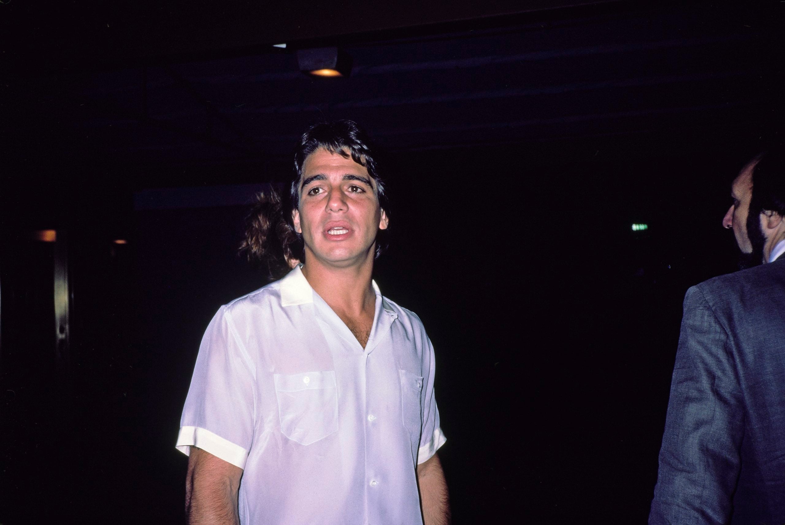 A man wearing a white short-sleeve shirt stands indoors in dim lighting. He has dark hair and is looking slightly off-camera. Another person in a suit is visible on the right, but only partially.