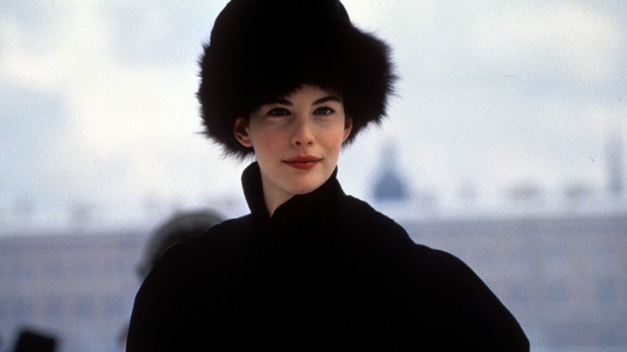 A woman wearing a large, furry black hat and a dark coat stands outdoors against a blurred, snowy city background.