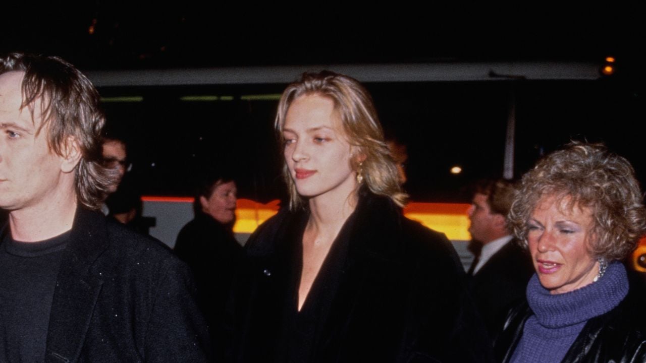 Three people walk outside at night, wearing dark clothing. The person in the center has shoulder-length blonde hair and a black coat. The people on either side have lighter hair; the right person wears a purple turtleneck.